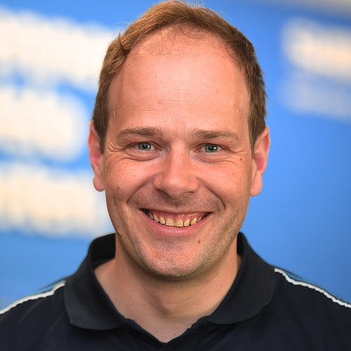 Lächelnder Mann mit kurzem Haar und einem poloähnlichen Shirt vor einem blauen Hintergrund.