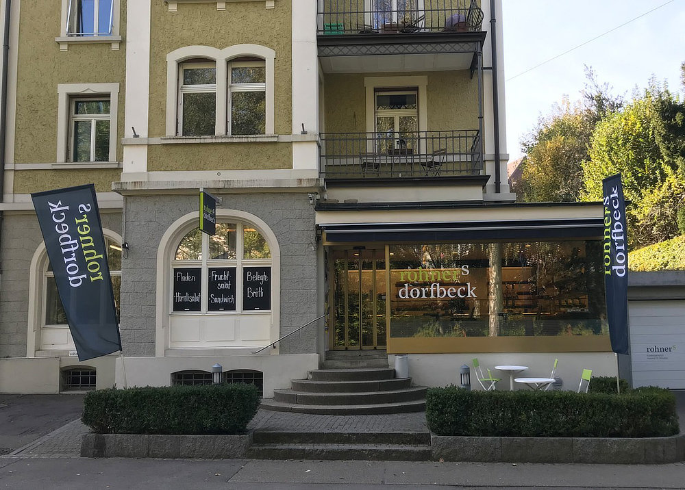 Die Fassade eines Restaurants mit der Beschriftung "rohner's dorfbeck" und einem einladenden Eingangsbereich, umgeben von Pflanzen und Fahnen.
