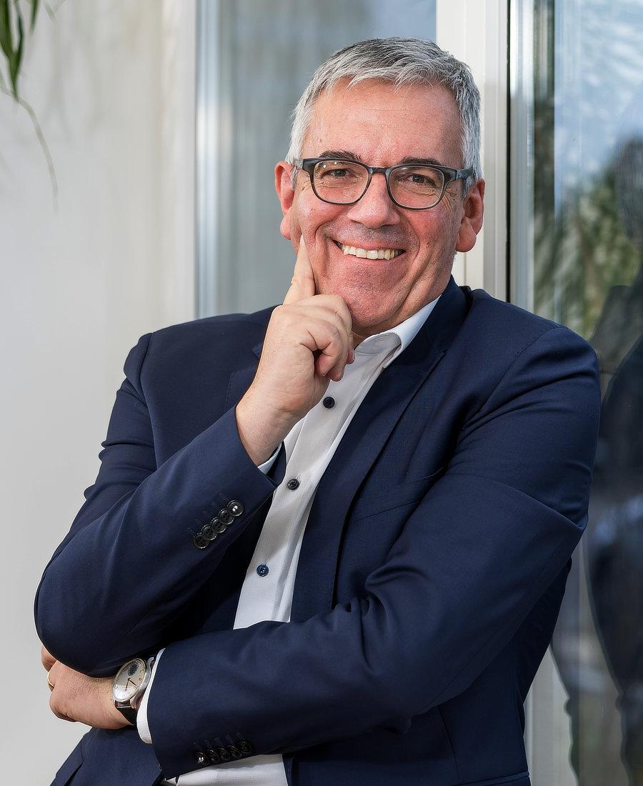 Ein lächelnder Mann mit Brille, der in einem blauen Anzug gekleidet ist, sitzt mit einem nachdenklichen Ausdruck und dem Finger an der Wange neben einem Fenster.